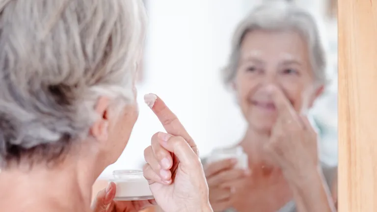 Older person looking into a mirror, applying cream to their nose while holding a jar.