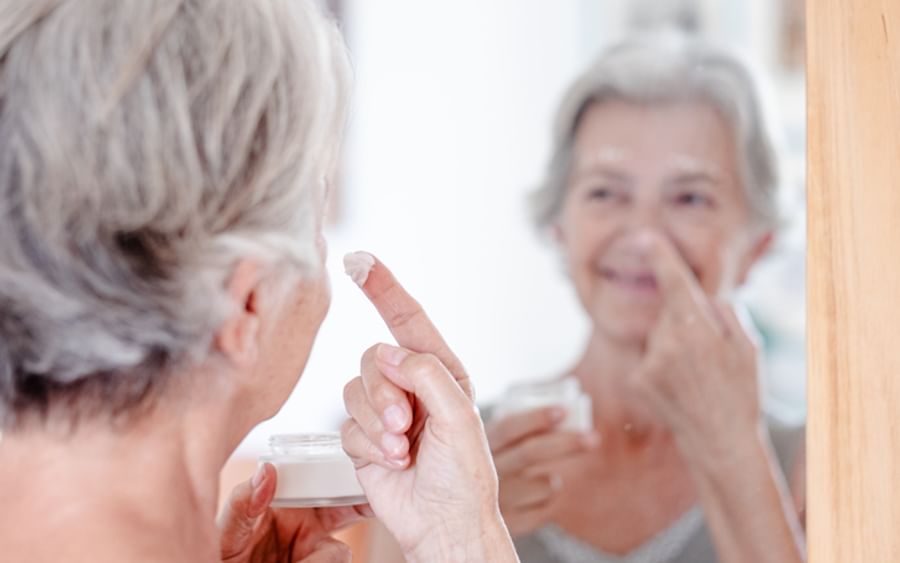 Older person looking into a mirror, applying cream to their nose while holding a jar.