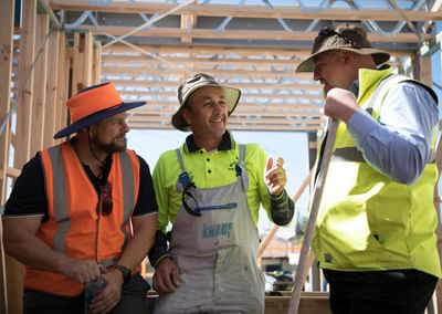 Three construction workers in hi-vis gear inside a wooden-framed building under construction.