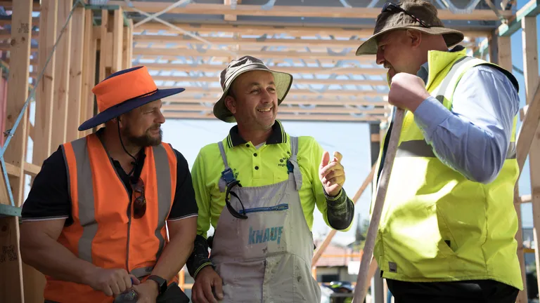 Three construction workers in hi-vis gear inside a wooden-framed building under construction.