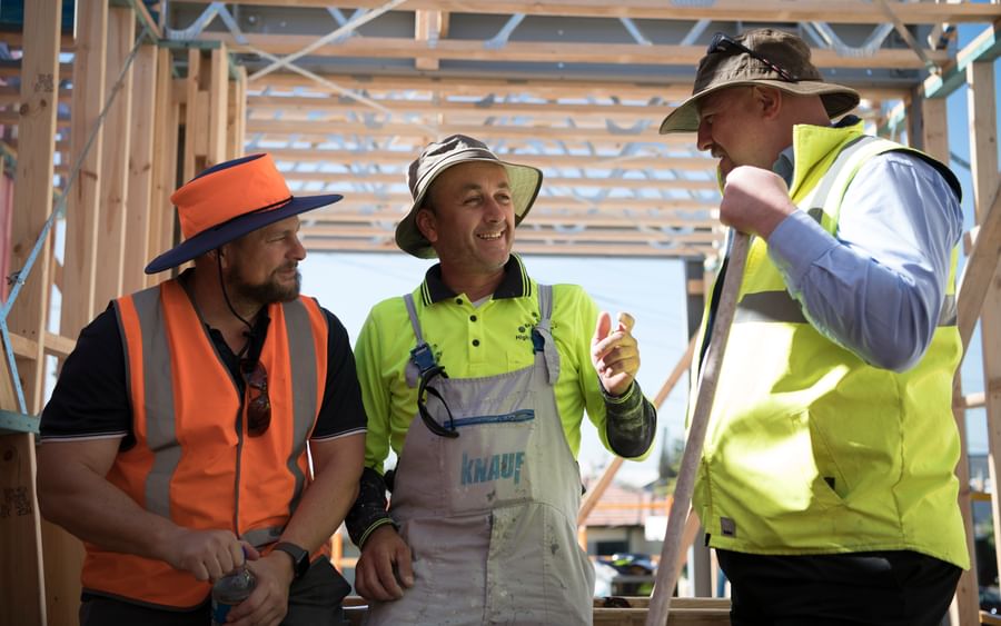 Three construction workers in hi-vis gear inside a wooden-framed building under construction.