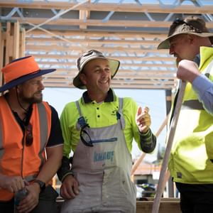 Three construction workers in hi-vis gear inside a wooden-framed building under construction.