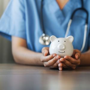 Healthcare worker in blue scrubs holds a white piggy bank on a wooden table, stethoscope around neck.