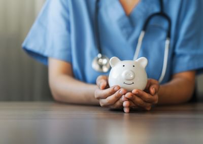Healthcare worker in blue scrubs holds a white piggy bank on a wooden table, stethoscope around neck.