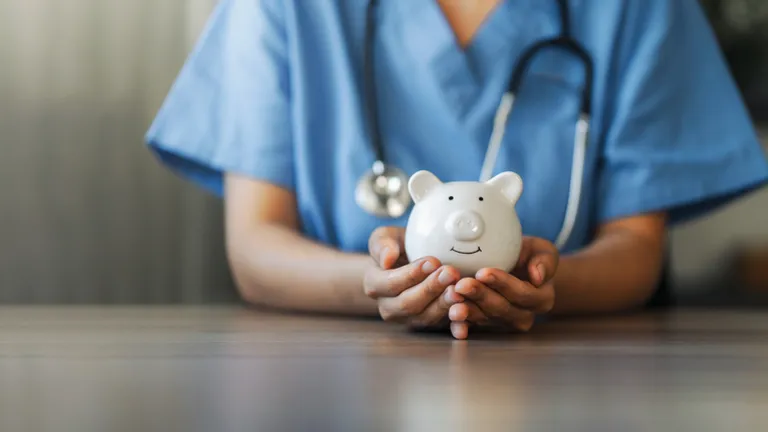 Healthcare worker in blue scrubs holds a white piggy bank on a wooden table, stethoscope around neck.