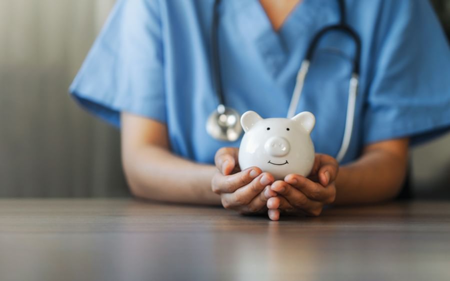 Healthcare worker in blue scrubs holds a white piggy bank on a wooden table, stethoscope around neck.