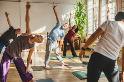 Group of people in a yoga class stretching with arms raised in a sunlit studio.