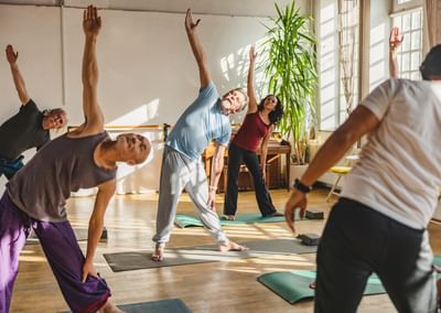 Group of people in a yoga class stretching with arms raised in a sunlit studio.