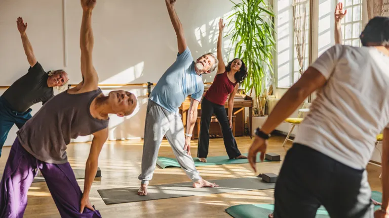 Group of people in a yoga class stretching with arms raised in a sunlit studio.