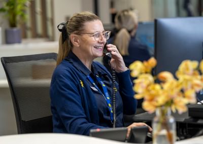 Smiling person with glasses at desk in office, talking on a corded phone, wearing a blue cardigan.