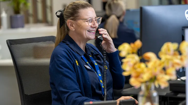 Smiling person with glasses at desk in office, talking on a corded phone, wearing a blue cardigan.