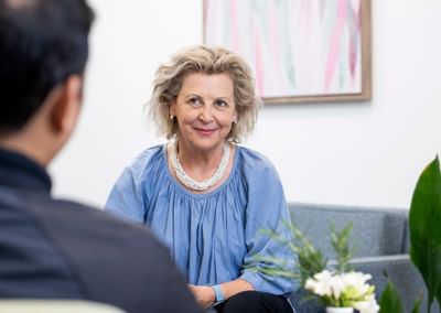 Smiling person with light hair in a blue top and white necklace, talking with someone.