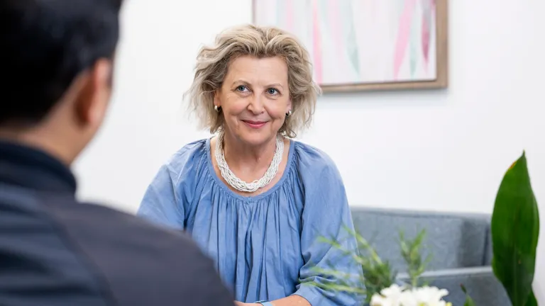 Smiling person with light hair in a blue top and white necklace, talking with someone.