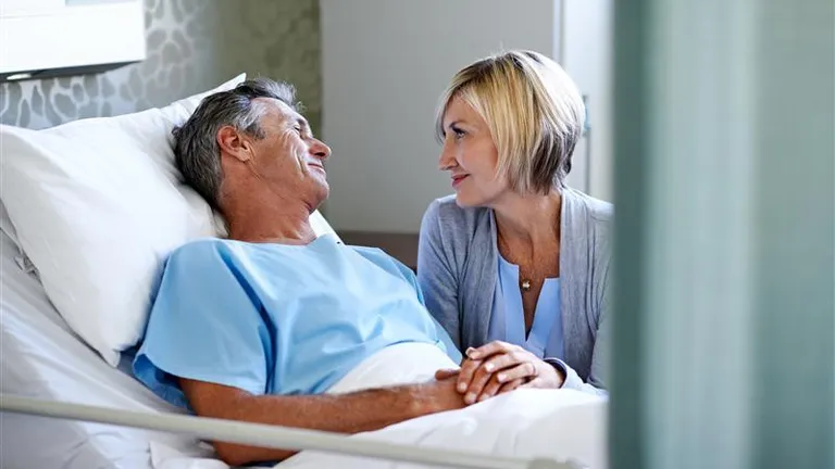 Older patient in a hospital bed wearing a blue gown, holding hands with a visitor.
