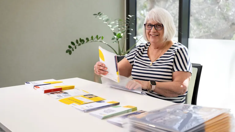 Older person with short white hair and glasses, in a striped shirt, at a table with brochures.