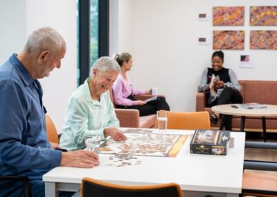 Two older adults piece together a jigsaw at a white table; puzzle box and glasses beside.