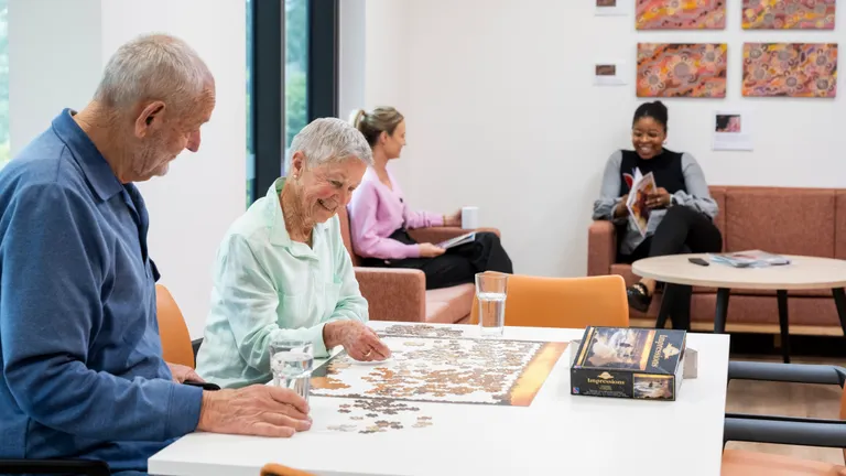 Two older adults piece together a jigsaw at a white table; puzzle box and glasses beside.