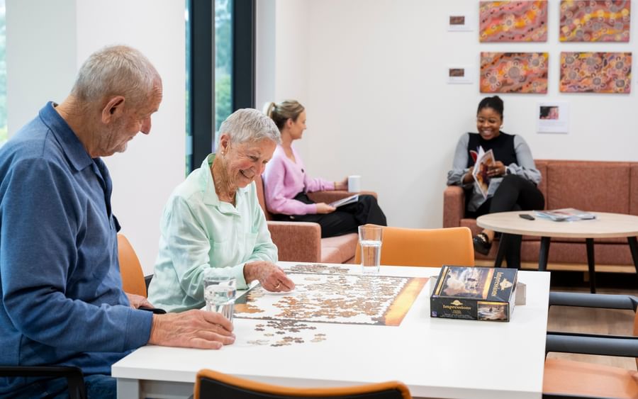 Two older adults piece together a jigsaw at a white table; puzzle box and glasses beside.