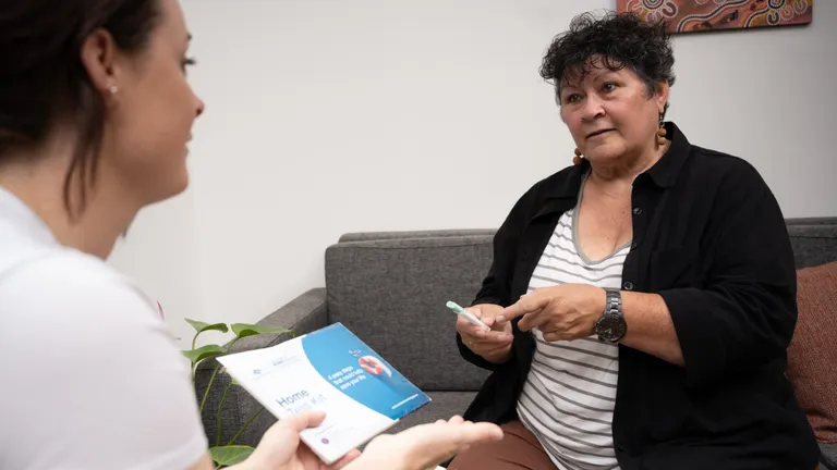 Two people sit on a grey sofa in a living room; one holds a brochure and points as they chat.