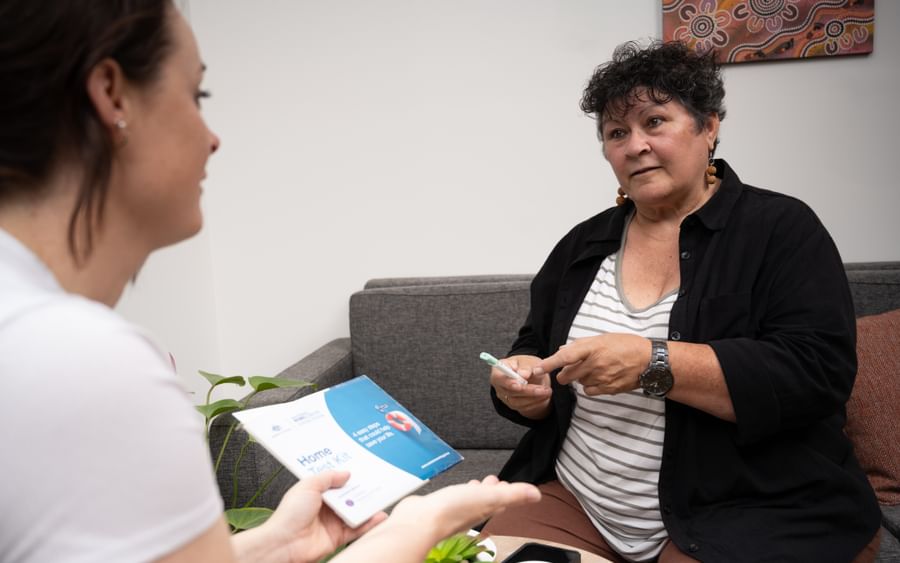 Two people sit on a grey sofa in a living room; one holds a brochure and points as they chat.
