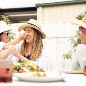 Two adults and a child wearing sun hats enjoy an outdoor meal at a table with salads and drinks.