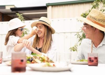 Two adults and a child wearing sun hats enjoy an outdoor meal at a table with salads and drinks.