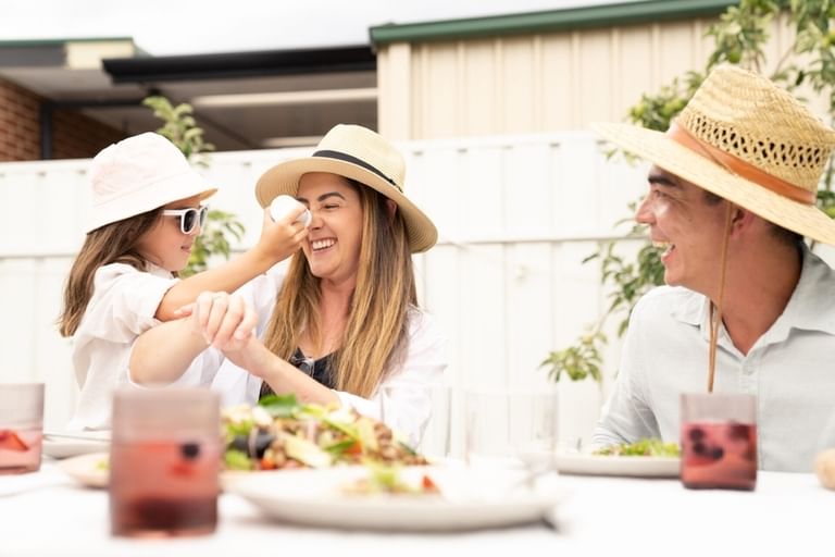 Two adults and a child wearing sun hats enjoy an outdoor meal at a table with salads and drinks.