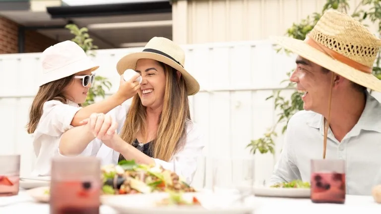 Two adults and a child wearing sun hats enjoy an outdoor meal at a table with salads and drinks.