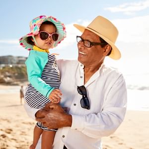 A person in a white shirt and straw hat holds a child on a sunny beach, both wearing sunglasses.