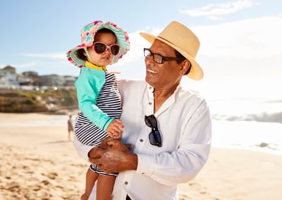 A person in a white shirt and straw hat holds a child on a sunny beach, both wearing sunglasses.