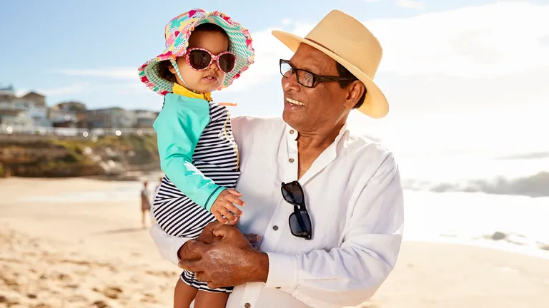 A person in a white shirt and straw hat holds a child on a sunny beach, both wearing sunglasses.