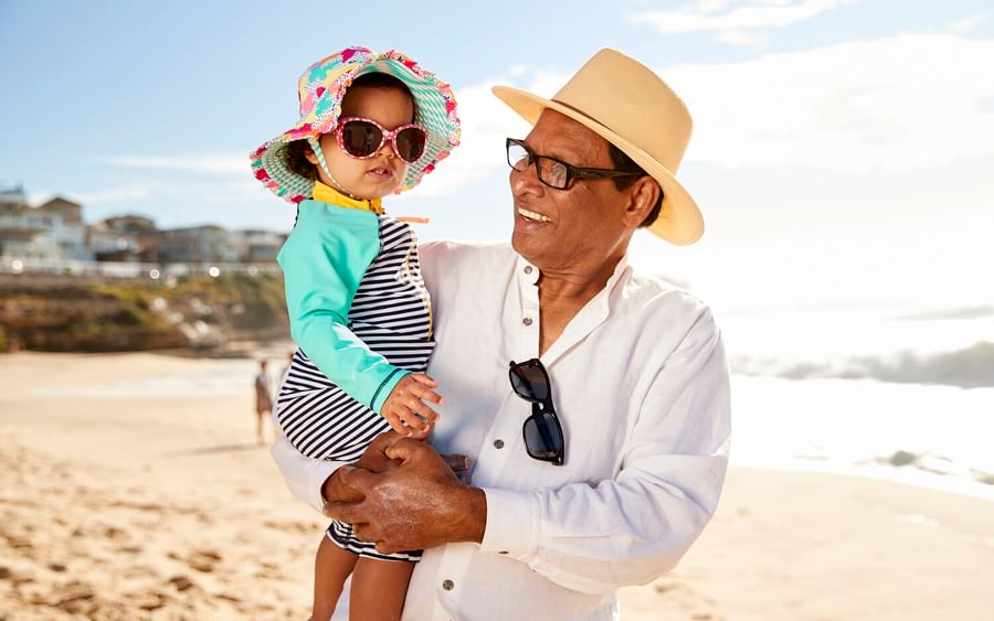 A person in a white shirt and straw hat holds a child on a sunny beach, both wearing sunglasses.