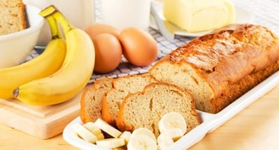 Banana bread loaf with sliced pieces on a white dish; bananas, eggs and butter in background.