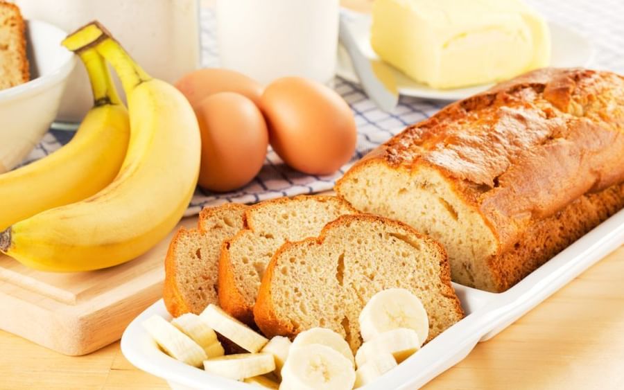 Banana bread loaf with sliced pieces on a white dish; bananas, eggs and butter in background.