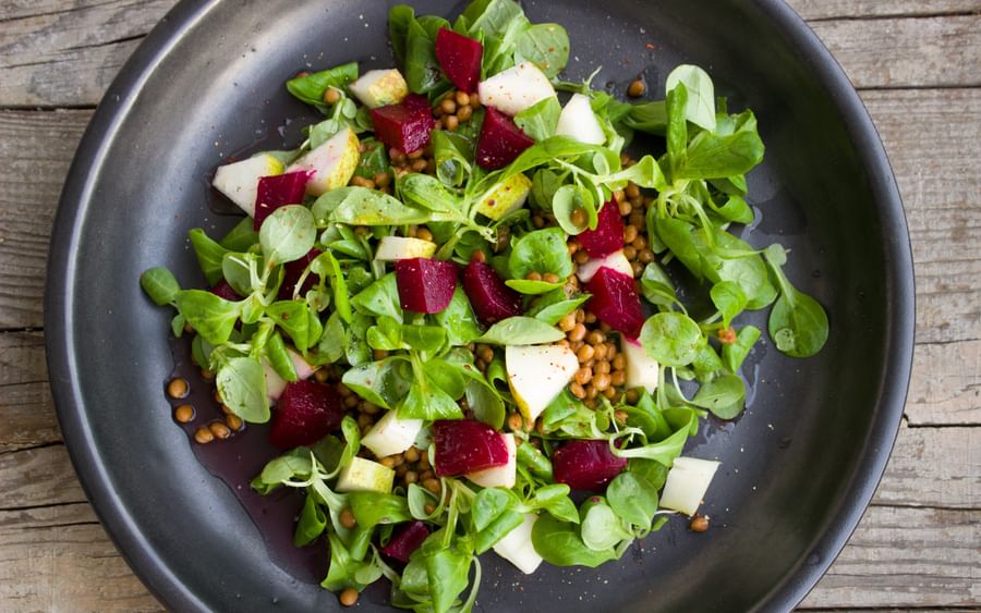 Dark plate with a green salad of beets, apple chunks and lentils on a wooden table.