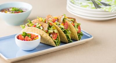 Four veggie tacos with lettuce, tomatoes, corn and peppers on a blue plate, with a salsa bowl nearby.