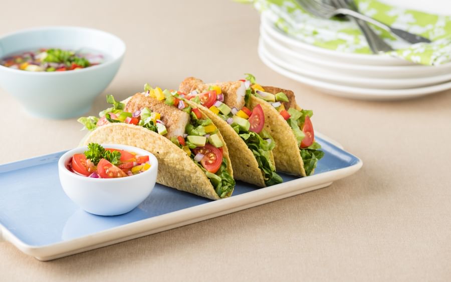 Four veggie tacos with lettuce, tomatoes, corn and peppers on a blue plate, with a salsa bowl nearby.