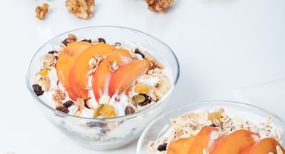 Two glass bowls of yogurt with granola, apricot slices, and dried fruits on a white surface.