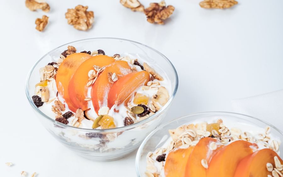 Two glass bowls of yogurt with granola, apricot slices, and dried fruits on a white surface.