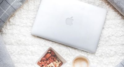 Cozy flat lay with a silver MacBook, granola bowl, coffee, and grey cushions on a white fluffy rug.