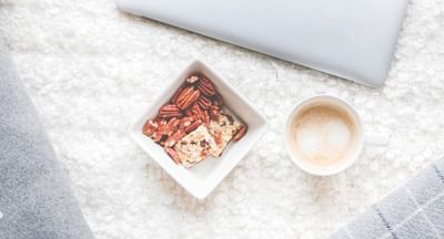 Cozy flat lay with a silver MacBook, granola bowl, coffee, and grey cushions on a white fluffy rug.