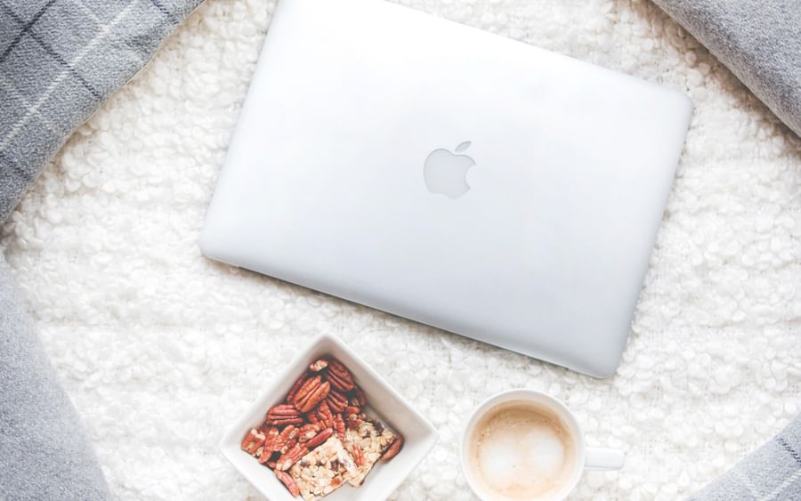 Cozy flat lay with a silver MacBook, granola bowl, coffee, and grey cushions on a white fluffy rug.