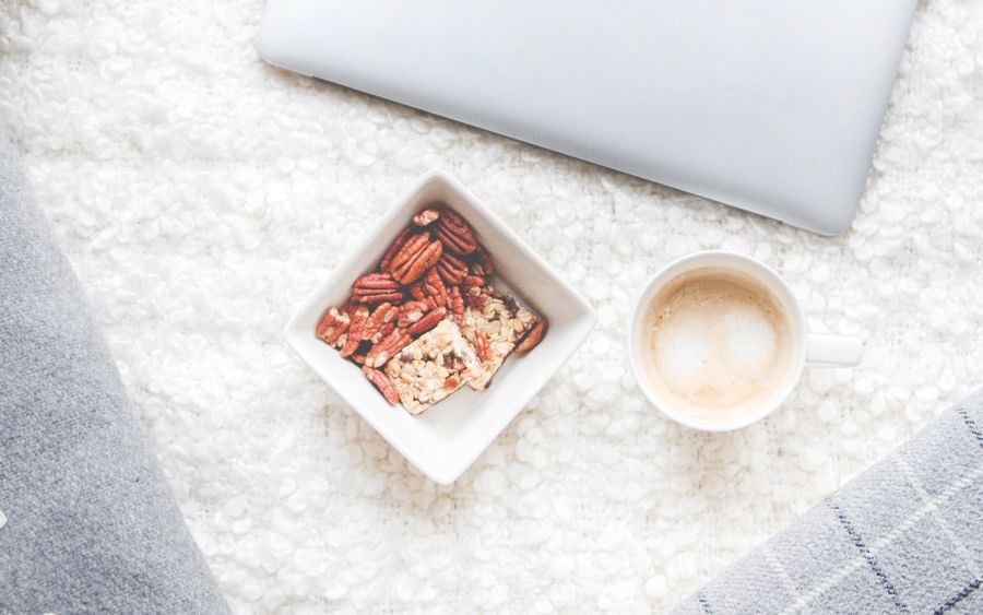 Cozy flat lay with a silver MacBook, granola bowl, coffee, and grey cushions on a white fluffy rug.