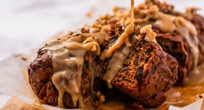 Caramel being poured over a nut-topped loaf cake on parchment, placed on a wooden board.