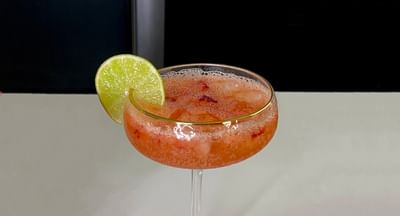 Pink mocktail with ice in a coupe glass, lime wedge garnish, on a light bar counter.
