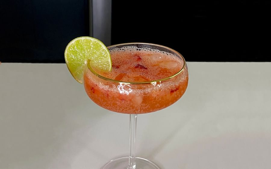 Pink mocktail with ice in a coupe glass, lime wedge garnish, on a light bar counter.