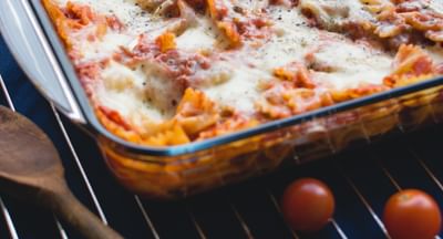 Baked pasta in a glass dish with melted cheese and tomato sauce; cherry tomatoes on a rack nearby.
