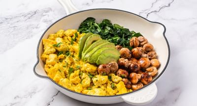 White frying pan on a marble surface with scrambled eggs, avocado slices, greens and mushrooms.