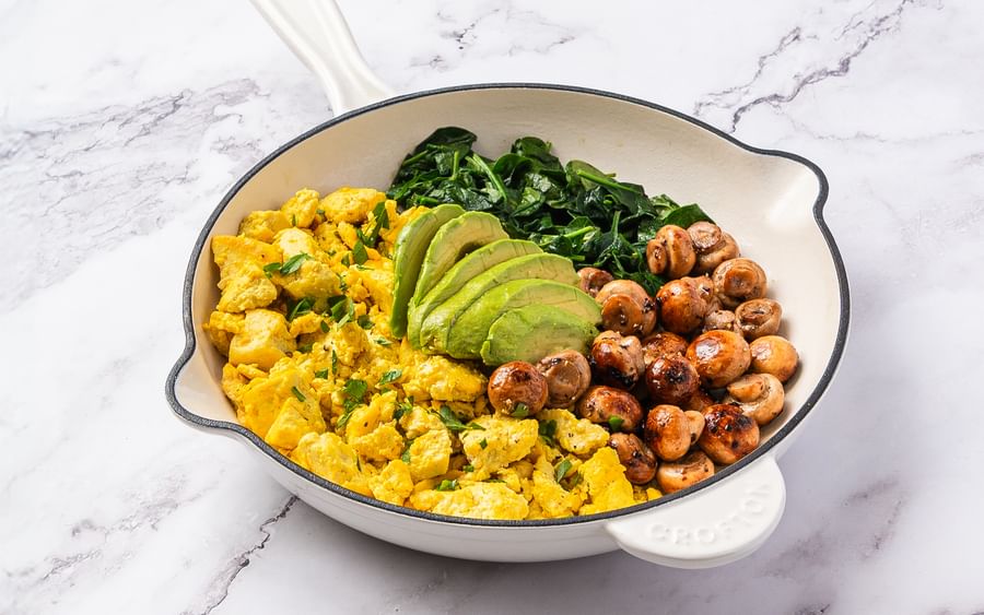 White frying pan on a marble surface with scrambled eggs, avocado slices, greens and mushrooms.
