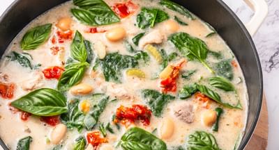Creamy white bean and spinach soup in a pot with basil, tomatoes, and bread slices nearby.